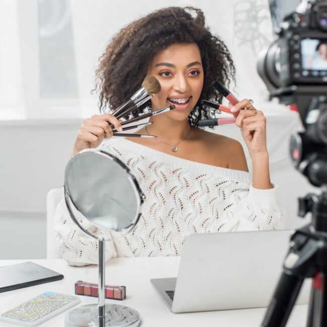 selective focus of happy african american influencer in braces holding lip glosses and cosmetic